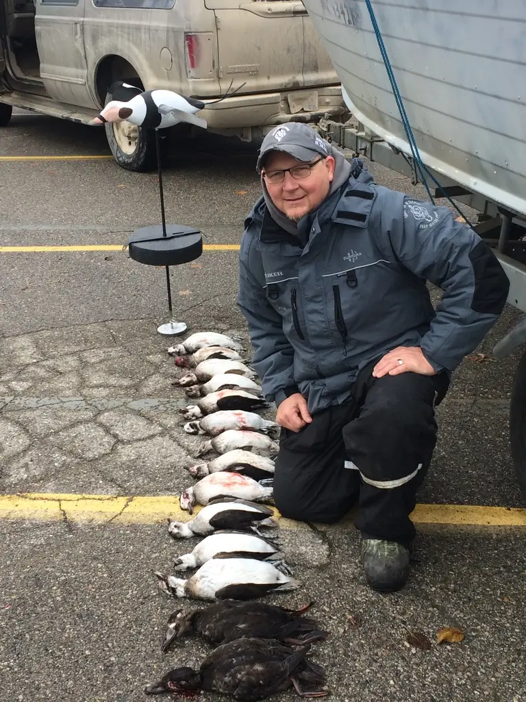 Jamie Hurlburt, the owner of Deep Water Duck, kneels in outdoor gear next to a row of about a dozen of his duck decoys laid out on the ground by a parked vehicle and a boat; a bird decoy stands on a pole behind him.
