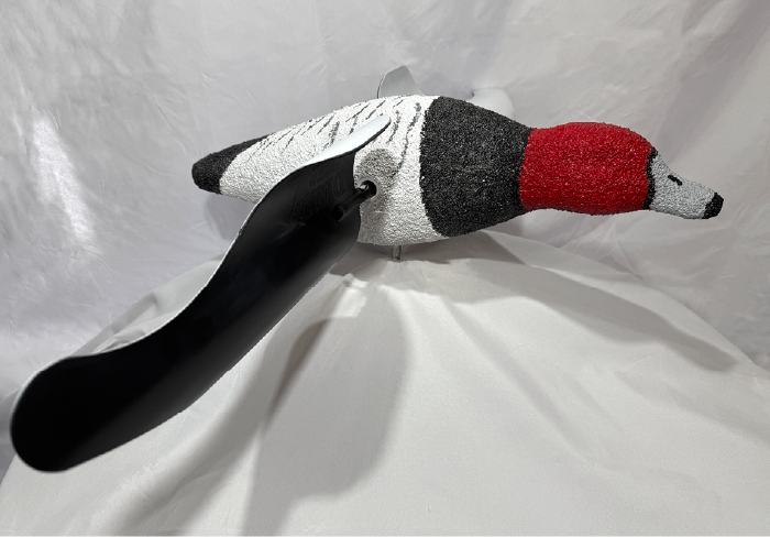 A bird-shaped decoy with a red head, black beak, and white and gray body is mounted on a metal stand. Its black plastic wing is extended, and the background is a white, draped fabric.
