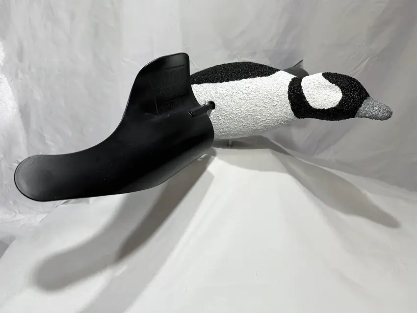 A realistic bufflehead duck decoy with a textured white body and a large, black plastic flipper attached to its side is displayed on a white fabric backdrop.