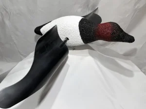 A realistic canvasback decoy duck with a white body, black chest, and reddish-brown head is displayed on a white fabric backdrop.