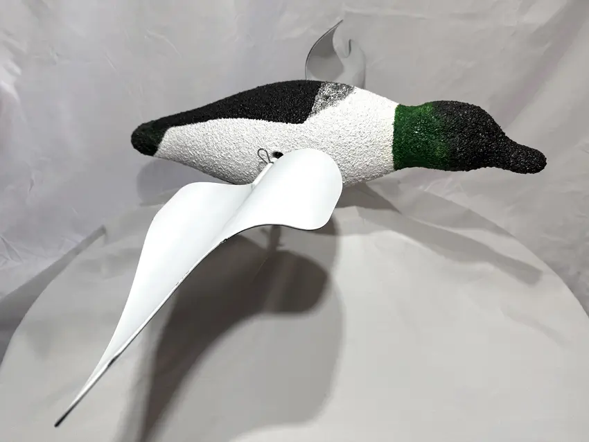 A realistic spoony duck decoy with a green head and a textured black-and-white body, and is displayed on a white fabric backdrop.