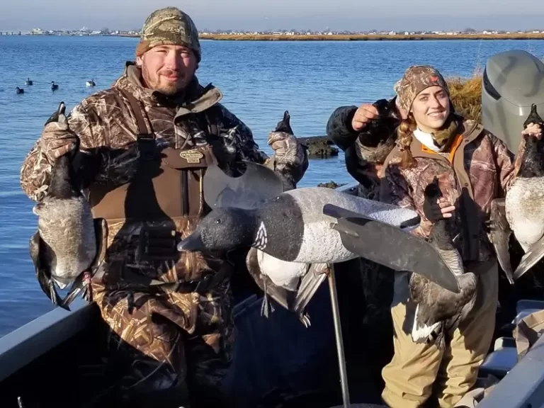 Two people in camouflage hunting gear stand on a boat by the water, holding up dead ducks surrounded by duck decoys.
