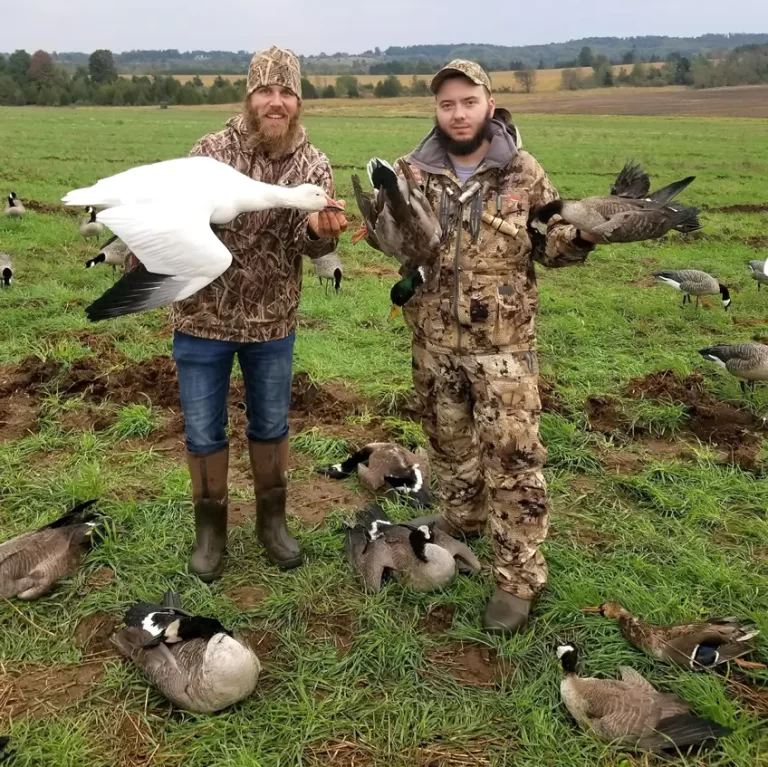 Two men in camouflage gear stand in a grassy field holding ducks surrounded by more ducks on the ground.