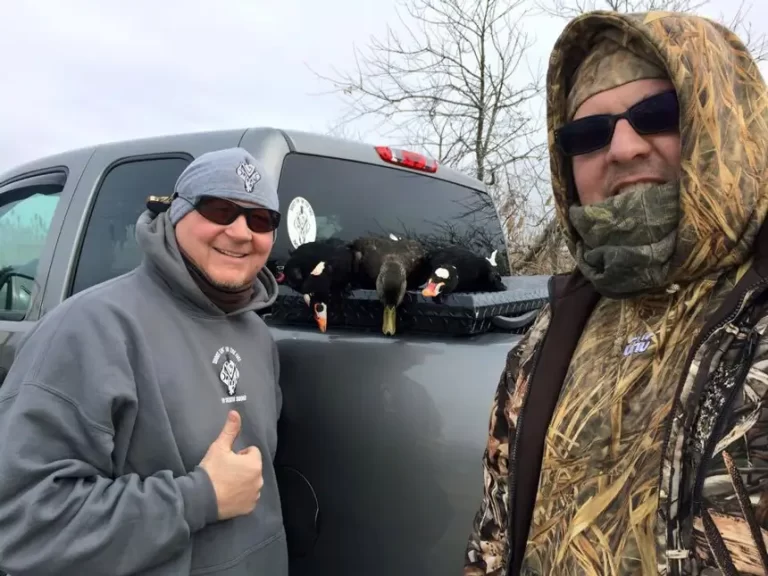 Two men in camouflage and outdoor gear pose in front of a truck with three hunted ducks laid out on the truck bed.