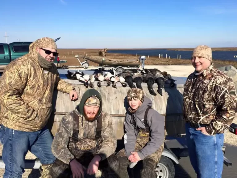 Four men in camouflage and outdoor gear pose in front of a trailer with several hunted birds laid out on top.