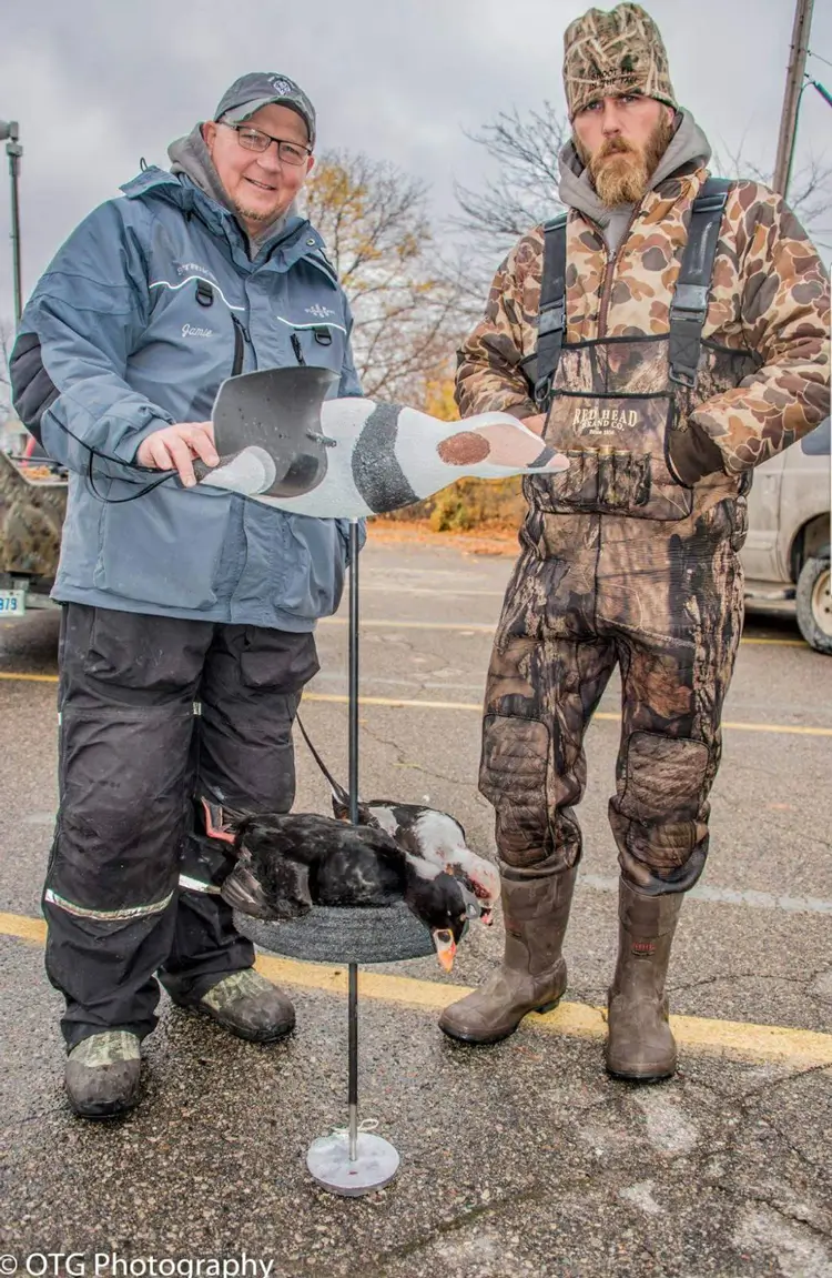 Two men in outdoor gear stand in a parking lot posing next to a display of realistic floating duck decoy with dead ducks underneath.