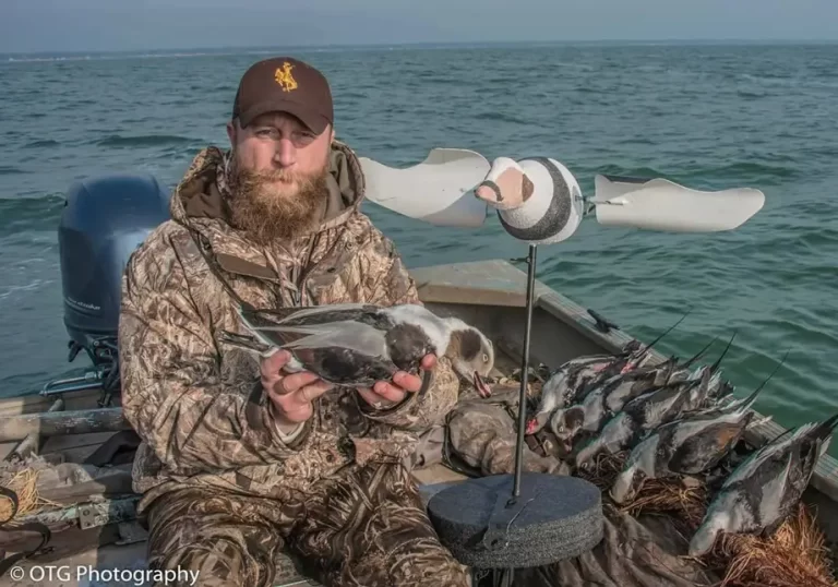 A man in camouflage gear sits in a boat holding a dead duck surrounded by more ducks and a floating duck decoy on open waters.