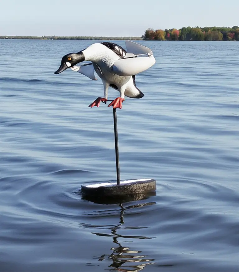 A realistic duck decoy mounted on a pole stands above the surface of a calm lake, with gentle ripples in the water and a tree-lined shoreline visible in the background.